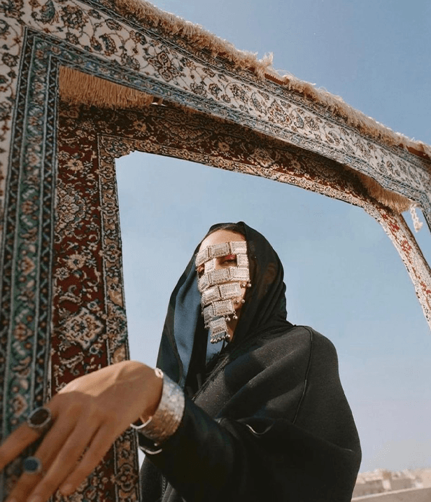 veiled figure framed by a hanging persian carpet, gulf sky behind